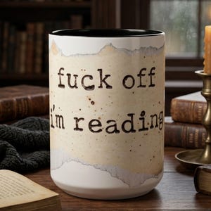 Black accent ceramic reading mug with “Fuck Off I’m Reading” design on a wooden table surrounded by vintage books, a candle and a dark knit blanket in a cozy rainy library setting.