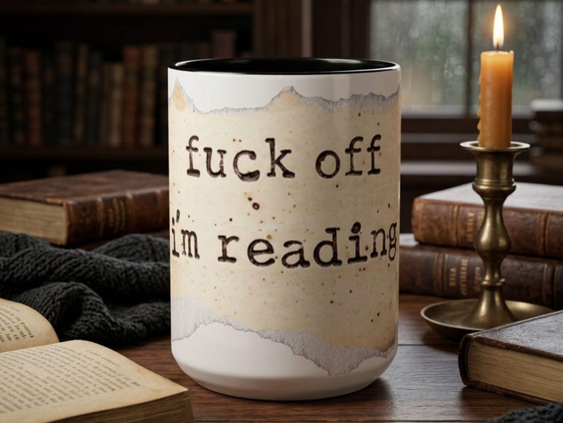 Black accent ceramic reading mug with “Fuck Off I’m Reading” design on a wooden table surrounded by vintage books, a candle and a dark knit blanket in a cozy rainy library setting.