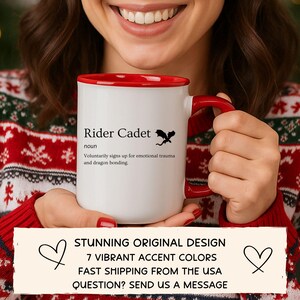 May include: A white ceramic mug with a red interior and handle, featuring the text "Rider Cadet" and a dragon silhouette. The mug also has the definition of "Rider Cadet". The mug is held by a person wearing a red and white sweater.