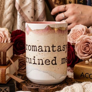 May include: A ceramic mug with the text "romantasy ruined me" in black lettering. The mug has a white base with a watercolor design in shades of pink, beige, and gold. Roses and books are in the background.