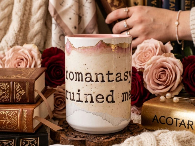 May include: A ceramic mug with the text "romantasy ruined me" in black lettering. The mug has a white base with a watercolor design in shades of pink, beige, and gold. Roses and books are in the background.