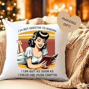 Op de afbeelding: Vierkant wit kussen met een retro illustratie van een lezende vrouw. De tekst op het kussen luidt "I'M NOT ADDICTED TO READING I CAN QUIT AS SOON AS I FINISH ONE MORE CHAPTER". Een hartvormig label zegt "AMAZING GIFT!"