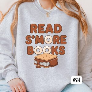 以下が含まれることがあります： 茶色の文字で「READ S'MORE BOOKS」と書かれた、明るい灰色のスウェットシャツ。テキストの下には、溶けたチョコレートが入ったスモアのグラフィックがあります。右下には白いボックスに「ASH」という文字があります。