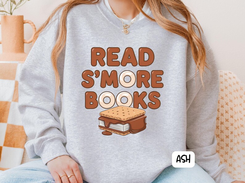 以下が含まれることがあります： 茶色の文字で「READ S'MORE BOOKS」と書かれた、明るい灰色のスウェットシャツ。テキストの下には、溶けたチョコレートが入ったスモアのグラフィックがあります。右下には白いボックスに「ASH」という文字があります。