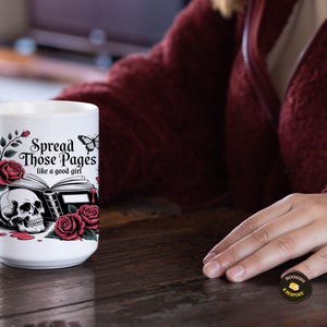 May include: White ceramic mug with a gothic design featuring a skull, roses, and the text "Spread Those Pages like a good girl." The mug has a black and red color scheme, with a butterfly accent.