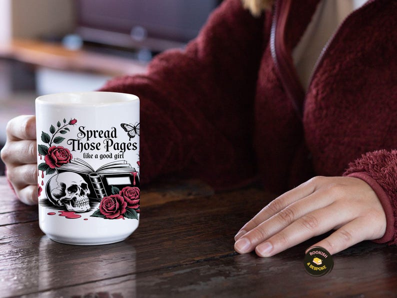 May include: White ceramic mug with a gothic design featuring a skull, roses, and the text "Spread Those Pages like a good girl." The mug has a black and red color scheme, with a butterfly accent.