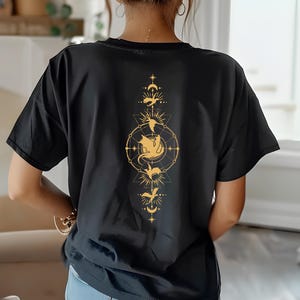 Puede incluir: Camiseta negra con un gr&aacute;fico dorado de un drag&oacute;n en un c&iacute;rculo con estrellas y lunas. El drag&oacute;n est&aacute; rodeado por un dise&ntilde;o geom&eacute;trico.