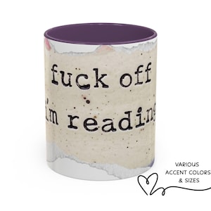 K&ouml;nnte beinhalten: Ein Keramikbecher mit violettem Innenraum und Rand, der eine beige, gesprenkelte, papierartige Textur aufweist. Der Text "fuck off i'm reading" ist in einer schwarzen Schriftart im Schreibmaschinenstil auf die Oberfl&auml;che gedruckt.