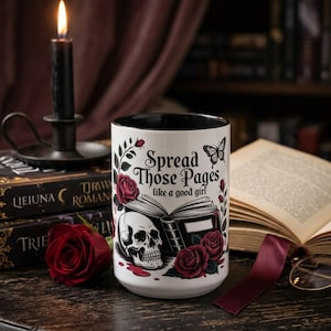 May include: White ceramic mug with a black interior, featuring the text "Spread Those Pages like a good girl" and illustrations of a skull, roses, and a butterfly. A lit black candle sits atop a stack of books.