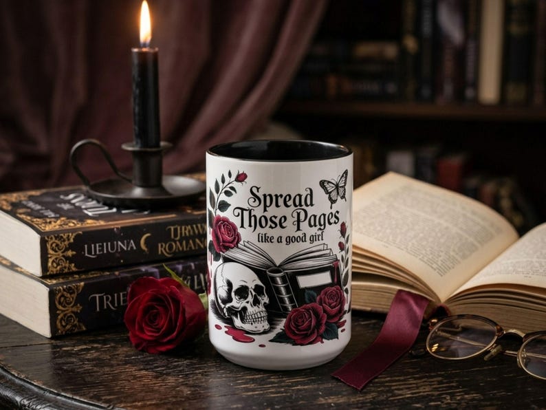 May include: White ceramic mug with a black interior, featuring the text "Spread Those Pages like a good girl" and illustrations of a skull, roses, and a butterfly. A lit black candle sits atop a stack of books.