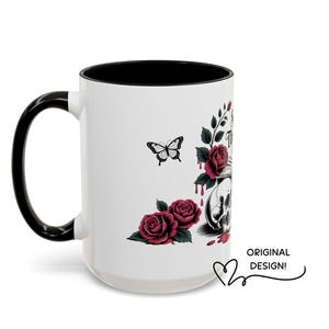 May include: White ceramic mug with a black handle and interior. The mug features a design of a skull, roses, and a butterfly. The words "ORIGINAL DESIGN!" are written on the mug.