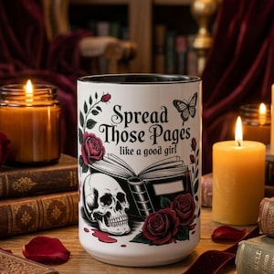 May include: A white ceramic mug with a black interior, featuring a skull, roses, and the text "Spread Those Pages like a good girl." The mug is surrounded by lit candles, books, and red rose petals.