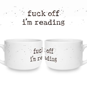 K&ouml;nnte beinhalten: Zwei wei&szlig;e Keramiktassen mit dem schwarzen, im Schreibmaschinenstil gedruckten Text "fuck off i'm reading" auf der Vorderseite. Das Design enth&auml;lt kleine schwarze Sprenkel um den Text herum, die ein abgenutztes Aussehen erzeugen.