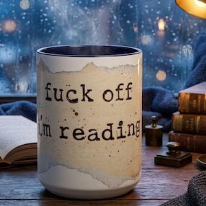 Navy accent ceramic reading mug with “Fuck Off I’m Reading” design on a wooden table surrounded by books, candlelight and a navy knit blanket in a cozy rainy nighttime reading nook.