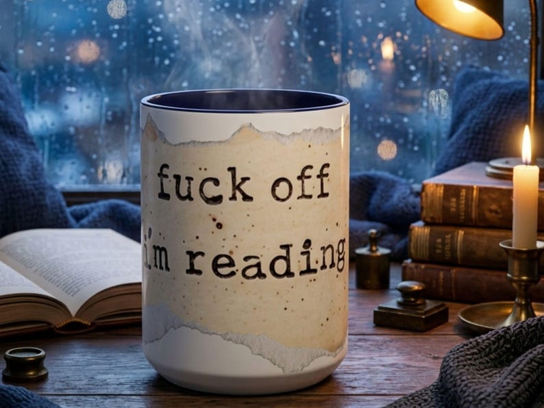 Navy accent ceramic reading mug with “Fuck Off I’m Reading” design on a wooden table surrounded by books, candlelight and a navy knit blanket in a cozy rainy nighttime reading nook.