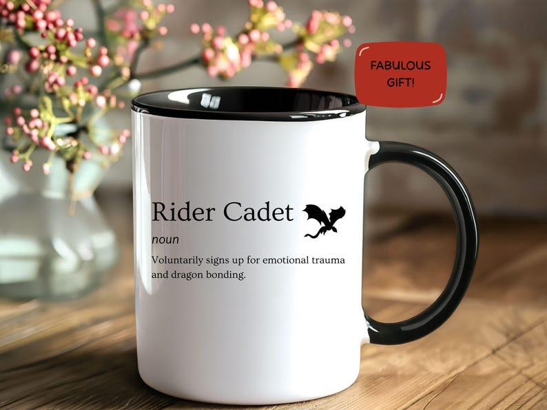 May include: White ceramic mug with a black handle and rim. The mug features the text "Rider Cadet" with a dragon graphic and the phrase "Voluntarily signs up for emotional trauma and dragon bonding." A red tag reads "FABULOUS GIFT!"