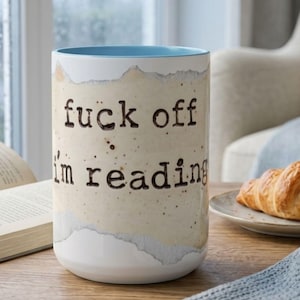 Light blue accent ceramic reading mug with “Fuck Off I’m Reading” design on a wooden table beside an open book, croissant and soft blue knit blanket in a bright cozy rainy reading nook.