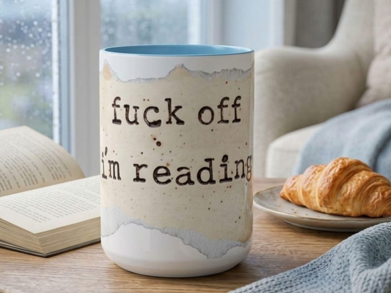 Light blue accent ceramic reading mug with “Fuck Off I’m Reading” design on a wooden table beside an open book, croissant and soft blue knit blanket in a bright cozy rainy reading nook.