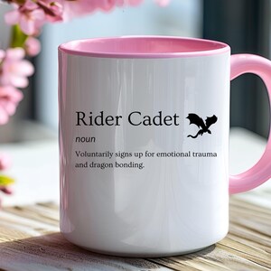 May include: White ceramic mug with a pink interior and handle. The mug features the text "Rider Cadet" with a definition and a black dragon silhouette. The mug is on a wooden surface with pink flowers in the background.
