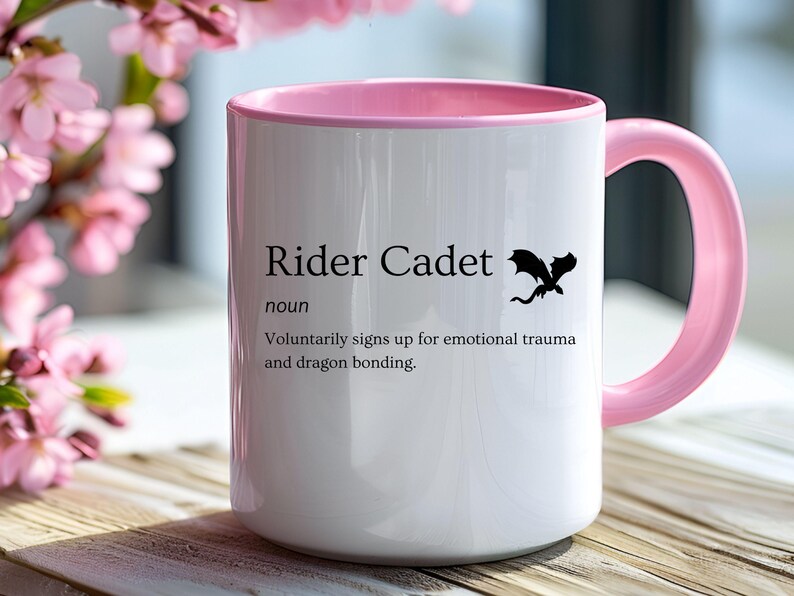 May include: White ceramic mug with a pink interior and handle. The mug features the text "Rider Cadet" with a definition and a black dragon silhouette. The mug is on a wooden surface with pink flowers in the background.