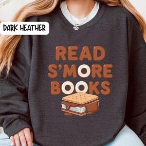 以下が含まれることがあります： 「READ S'MORE BOOKS」の文字が茶色と白でプリントされたダークヘザーのスウェットシャツ。テキストの下にはスモアのグラフィックがあります。スウェットシャツは濃いグレー色です。