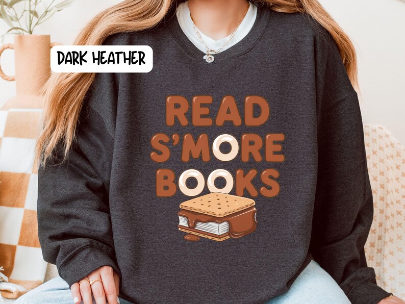 以下が含まれることがあります： 「READ S'MORE BOOKS」の文字が茶色と白でプリントされたダークヘザーのスウェットシャツ。テキストの下にはスモアのグラフィックがあります。スウェットシャツは濃いグレー色です。