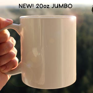 May include: A jumbo-sized, off-white ceramic mug with a curved handle. The mug is labeled "NEW! 20oz JUMBO" in black text. The mug is held by a hand, with a blurred green background.