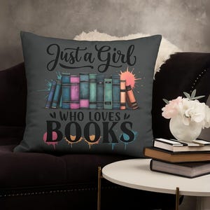 May include: A dark gray throw pillow with the text "Just a Girl Who Loves Books" and a colorful illustration of a stack of books. The pillow is on a dark brown couch.