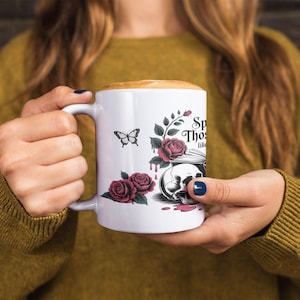 May include: White ceramic mug with a handle, featuring a butterfly, roses, and a skull design. The mug has the text "Speak Those Thoughts Like". The mug is filled with a coffee drink.