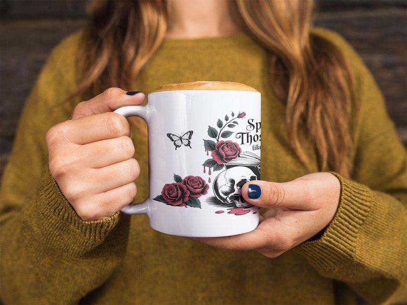 May include: White ceramic mug with a handle, featuring a butterfly, roses, and a skull design. The mug has the text "Speak Those Thoughts Like". The mug is filled with a coffee drink.