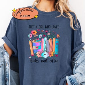 Puede incluir: Camiseta azul denim con un diseño colorido de libros, flores y una taza de café. El texto dice "Just a girl who loves books and coffee". La camiseta tiene un corte relajado y está hecha de un material suave.