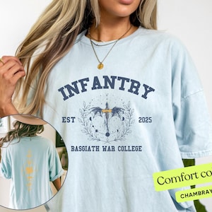 Puede incluir: Una camiseta azul claro con un estampado gr&aacute;fico de un drag&oacute;n y el texto "INFANTRY EST 2025 BASSGIATH WAR COLLEGE". La camiseta est&aacute; hecha de una tela suave y c&oacute;moda.