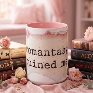 May include: A ceramic mug with the words "romantasy ruined me" in black lettering. The mug has a pink interior and a textured, off-white exterior with pink accents. The mug is surrounded by books and flowers.
