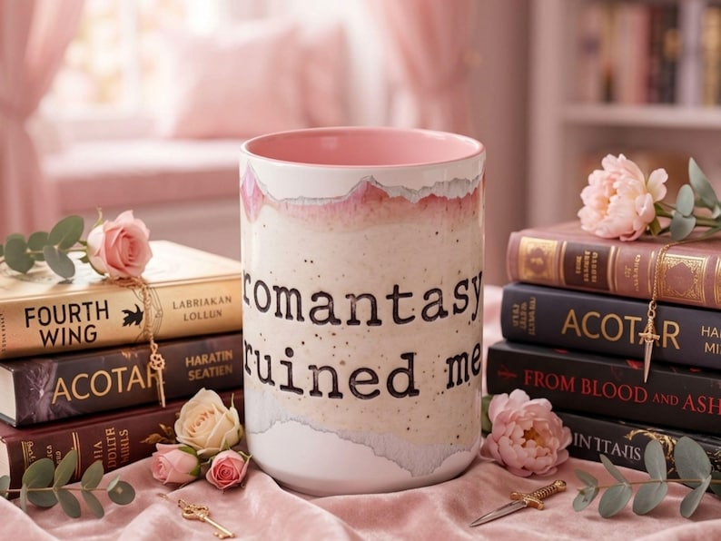 May include: A ceramic mug with the words "romantasy ruined me" in black lettering. The mug has a pink interior and a textured, off-white exterior with pink accents. The mug is surrounded by books and flowers.