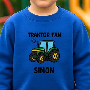 May include: A royal blue long-sleeve sweatshirt featuring a graphic of a green and yellow tractor with the text 'TRAKTOR-FAN' above and the name 'SIMON' below. The garment has a crew neckline and ribbed cuffs, with a small green icon indicating 80% cotton.
