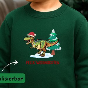 Könnte beinhalten: Ein dunkelgrünes Langarm-Sweatshirt mit einer Grafik eines Tyrannosaurus Rex mit Weihnachtsmütze neben einem Weihnachtsbaum. Der Text FELIX WEIHNACHTEN ist in Rot unter dem Dinosaurier aufgedruckt. Das Kleidungsstück hat einen Rundhalsausschnitt und gerippte Bündchen.