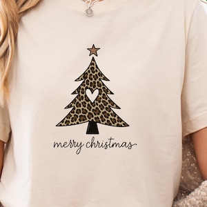 Könnte beinhalten: Beiges T-Shirt mit einem Leopardenmuster-Weihnachtsbaum-Design, einem Stern und einem Herz. Die Worte "merry christmas" sind darunter geschrieben. Das T-Shirt besteht aus 100% Baumwolle. Der Text "Leo-Look zu Weihnachten" ist ebenfalls vorhanden.