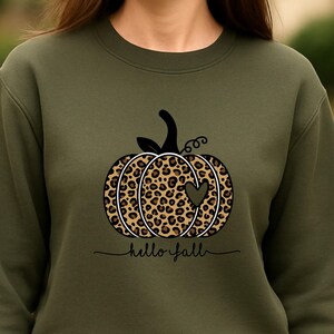Könnte beinhalten: Ein olivgrünes Sweatshirt mit einem Leopardenmuster-Kürbis-Design, einem Herz-Ausschnitt und dem Schriftzug "hello fall". Das Sweatshirt zeigt auch die Texte "50% Baumwolle" und "weitere Farben".