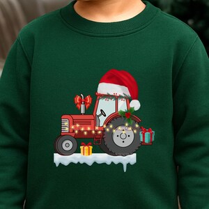 Könnte beinhalten: Ein dunkelgrünes Langarm-Sweatshirt mit einer Grafik eines roten Traktors, der eine Weihnachtsmütze trägt. Der Traktor ist mit Lichterketten, Stechpalmen und Geschenken verziert und steht auf einer weißen Schneefläche.