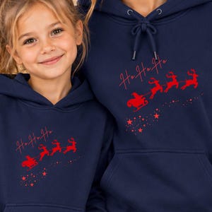 Könnte beinhalten: Zwei marineblaue Kapuzenpullover mit einem roten Grafikdruck von einem Weihnachtsmann im Schlitten, der von Rentieren gezogen wird, begleitet vom Text "Ho Ho Ho" und kleinen Sternen. Jeder Hoodie hat eine Kängurutasche und eine Kapuze.
