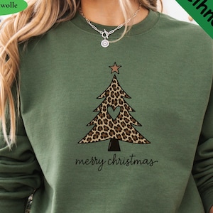 Könnte beinhalten: Ein olivgrüner Rundhals-Pullover mit einer Grafik eines Weihnachtsbaums im Leopardenmuster, einem kleinen herzförmigen Ausschnitt und einem Stern an der Spitze. Der Schriftzug "merry christmas" steht in schwarzer Schreibschrift darunter.