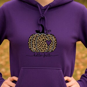 Könnte beinhalten: Ein lila Kapuzenpullover mit einem Kürbismotiv im Leopardenmuster und einem Herz. Unter dem Kürbis steht "hello fall". Der Hoodie hat eine Vordertasche und eine Kapuze mit Kordelzug.