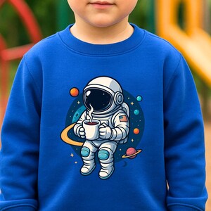 May include: A royal blue long-sleeve sweatshirt featuring a graphic of an astronaut holding a steaming mug in space, surrounded by planets and stars. The garment includes text labels reading 80% Baumwolle, F&uuml;r kleine Weltall-Fans, and weitere Farben.