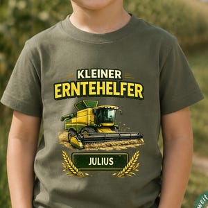 May include: An olive green short-sleeve t-shirt featuring a graphic of a yellow and green combine harvester. The text reads KLEINER ERNTEHELFER above the harvester and JULIUS inside a rectangular box below, flanked by two stalks of wheat.