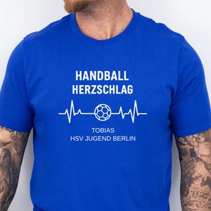 May include: A royal blue short-sleeved t-shirt featuring white text that reads HANDBALL HERZSCHLAG, a graphic of a handball within a heartbeat line, and the text TOBIAS HSV JUGEND BERLIN. The shirt also displays a small grey label with text.