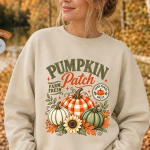 Könnte beinhalten: Ein beigefarbenes Sweatshirt mit Rundhalsausschnitt und einem Grafikdruck mit dem Text PUMPKIN Patch, FARM FRESH und HAND PICKED DAILY. Das Design zeigt einen großen orange-weiß karierten Kürbis, einen weißen Kürbis, einen grünen Kürbis und eine Sonnenblume.