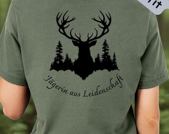 May include: Olive green t-shirt with a black silhouette of a deer and trees. The text "Jägerin aus Leidenschaft" is below the deer graphic. The shirt also has the text "Jägerinnen-Outfit" and "100% Baumwolle".