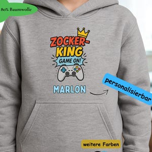 Könnte beinhalten: Ein grauer Kapuzenpullover mit Fronttasche und Kapuze. Auf der Vorderseite steht der Text "Zocker King" über einer Gamecontroller-Grafik und dem Namen "Marlon". Der Hoodie hat auch ein Etikett mit der Aufschrift "80% Baumwolle".
