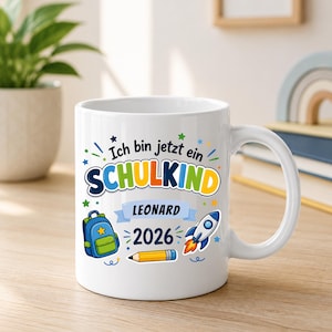 May include: A white ceramic mug featuring the German text Ich bin jetzt ein SCHULKIND, the name Leonard, and the year 2026. The design includes colorful illustrations of a blue backpack, a yellow pencil, and a rocket ship with orange flames.