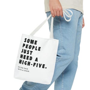Könnte beinhalten: Weiße Tragetasche mit schwarzem Text: „SOME PEOPLE JUST NEED A HIGH-FIVE. IN THE FACE. WITH A CHAIR.“ Die Tasche wird von einer Person mit hellblauen Jeans und einem hellen Oberteil getragen. Die Tasche hat weiße Träger.